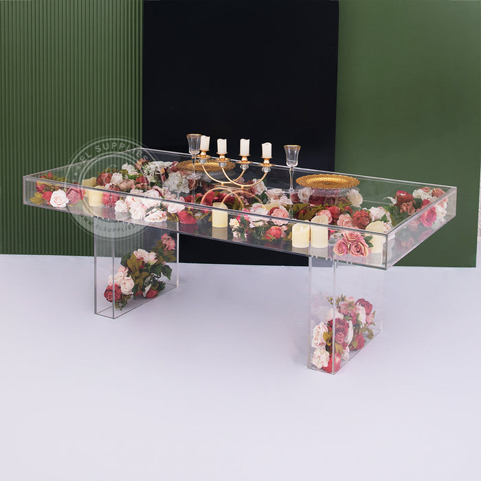 Clear acrylic dining table filled with flowers and candles inside, decorated with gold plates and a candle holder, placed in front of a green and black backdrop