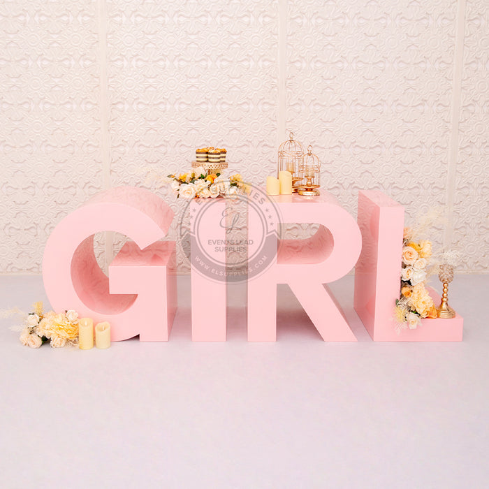 Large pink "GIRL" marquee letters decorated with flowers, candles and desserts, placed in front of a white patterned wall.