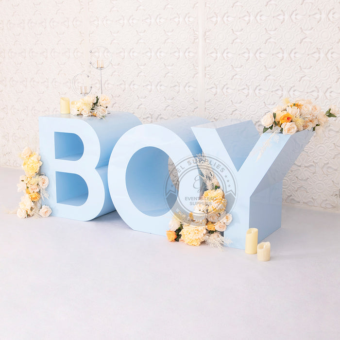 Large light blue "BOY" marquee letters decorated with flowers and candles, set against a white textured wall.
